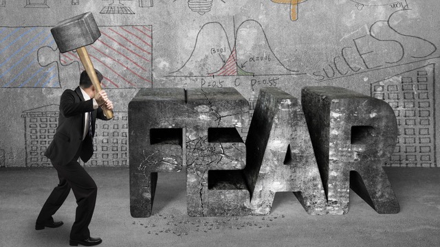 A person in a suit wielding a large hammer, poised to smash a concrete block spelling 'FEAR', representing the concept of overcoming beginner investor fears.