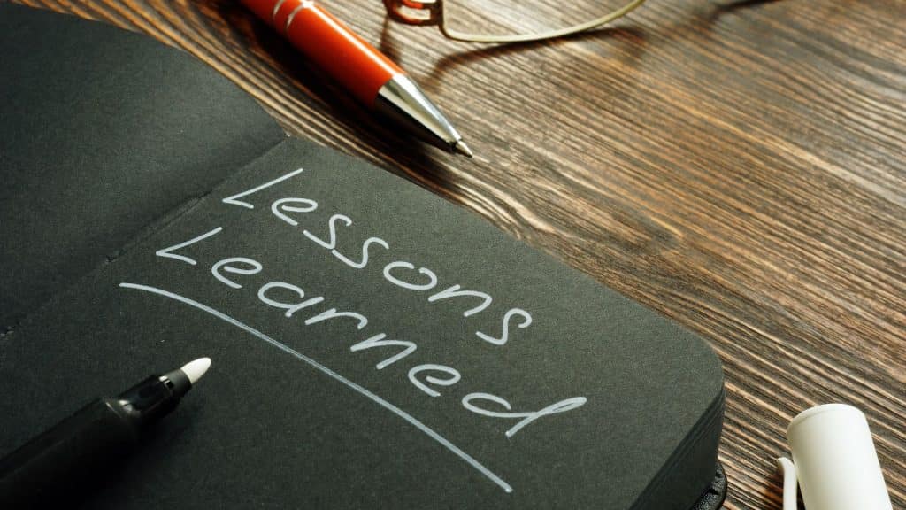 An open black notebook with 'Lessons Learned' written in white ink on the page, related to the Adele Dig interview. A red pen, eyeglasses, and a white marker are placed on a wooden desk nearby.