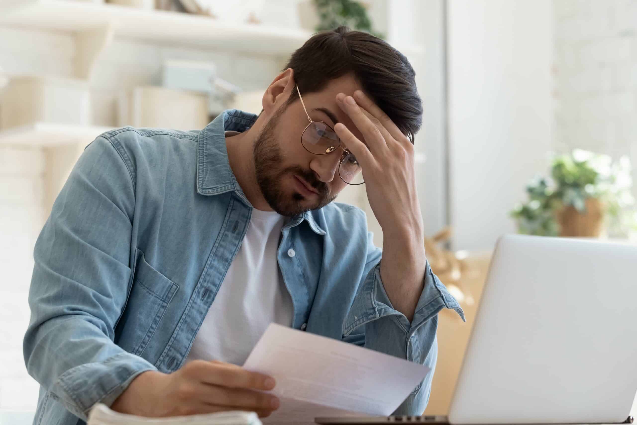 A stressed man in a denim shirt and glasses is holding his forehead while reading a document at a desk, likely dealing with issues related to problem tenants. His laptop is open in front of him, indicating that he might be researching or managing rental property concerns. This image relates to managing problem tenants effectively.
